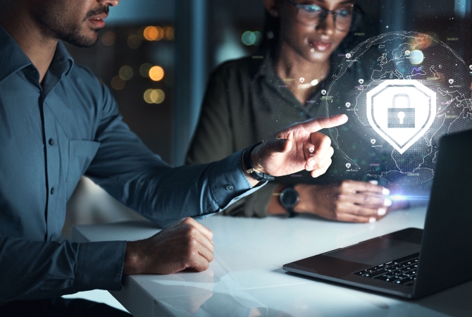 Two people sit at a desk with a laptop, one pointing at a digital padlock icon hologram, offering an overview of cybersecurity and data protection.